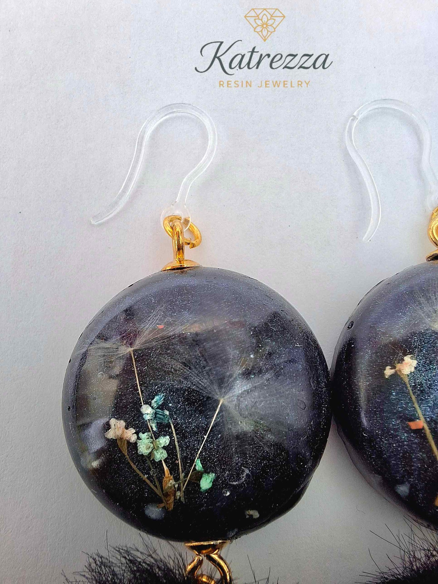 “Handmade black resin earrings with preserved flowers and dandelion seeds, accented with black pom-poms and gold details – hypoallergenic hooks.”