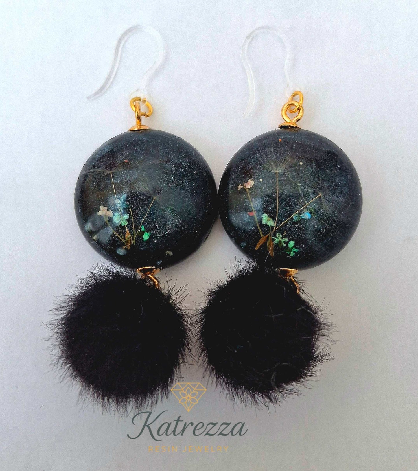 “Handmade black resin earrings with preserved flowers and dandelion seeds, accented with black pom-poms and gold details – hypoallergenic hooks.”