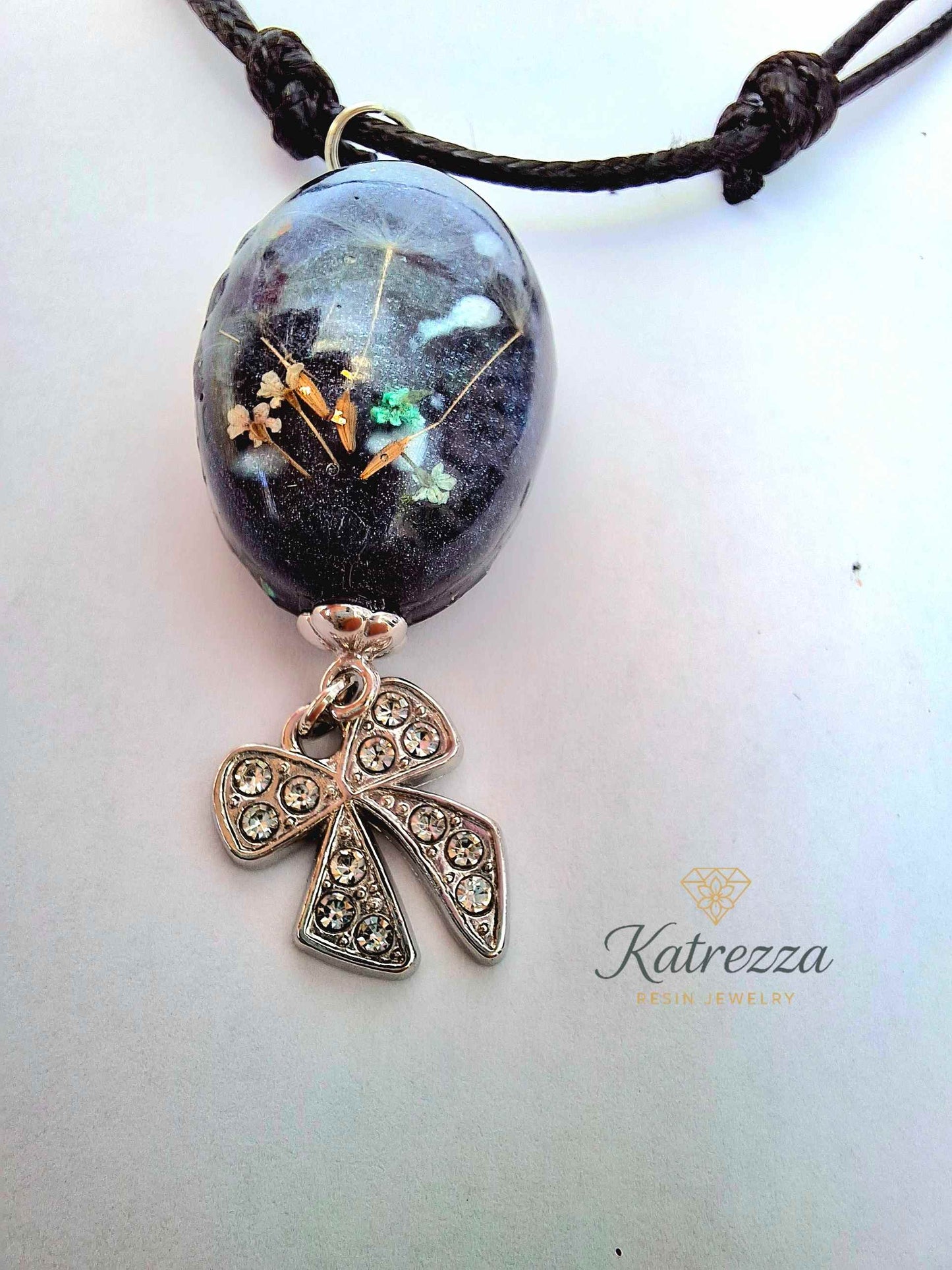 “Oval black resin pendant with real Queen Anne’s Lace flowers and dandelion seeds, finished with sparkling silver bow charm – Katrezza handmade jewelry.”
