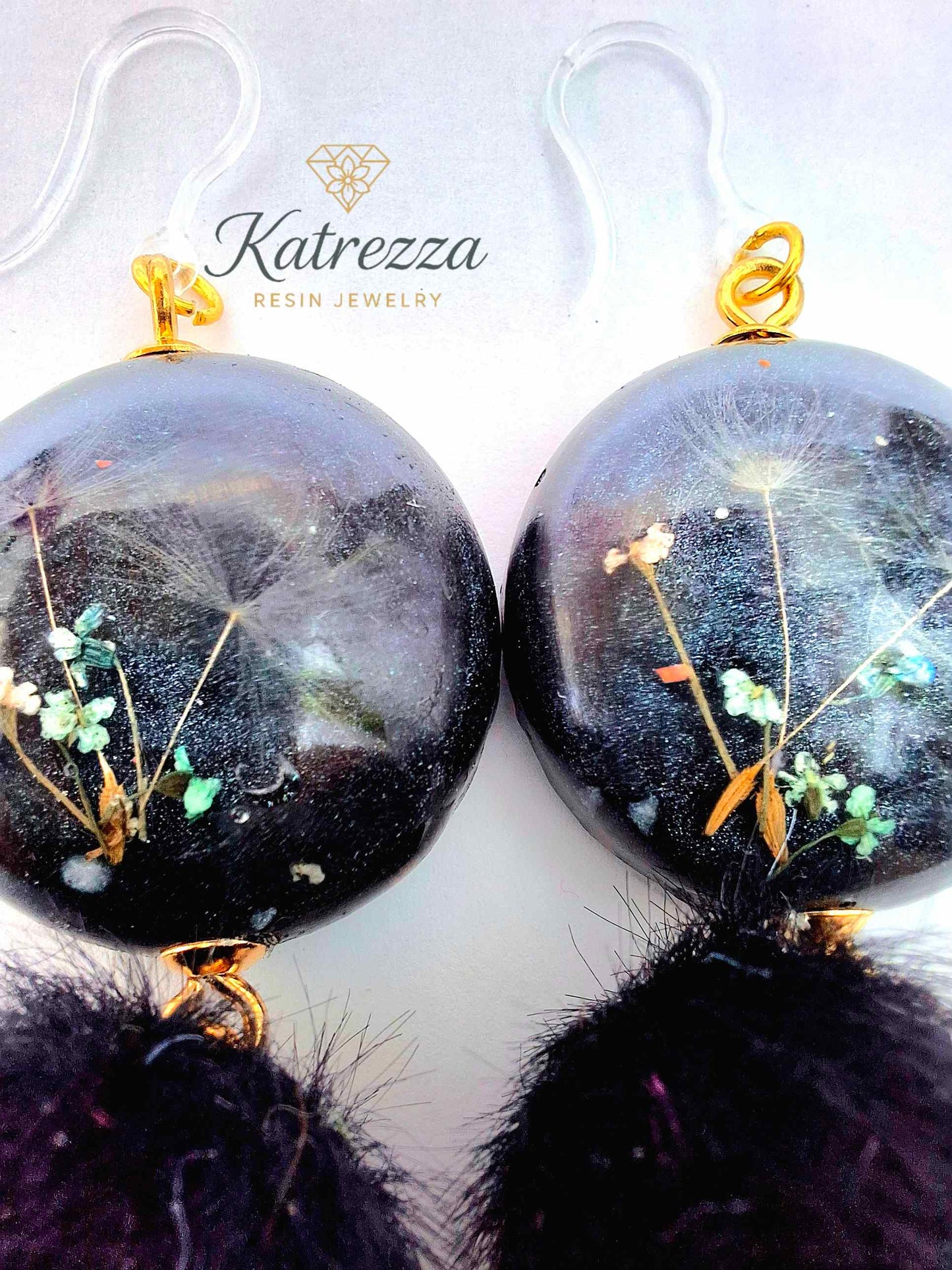 “Handmade black resin earrings with preserved flowers and dandelion seeds, accented with black pom-poms and gold details – hypoallergenic hooks.”