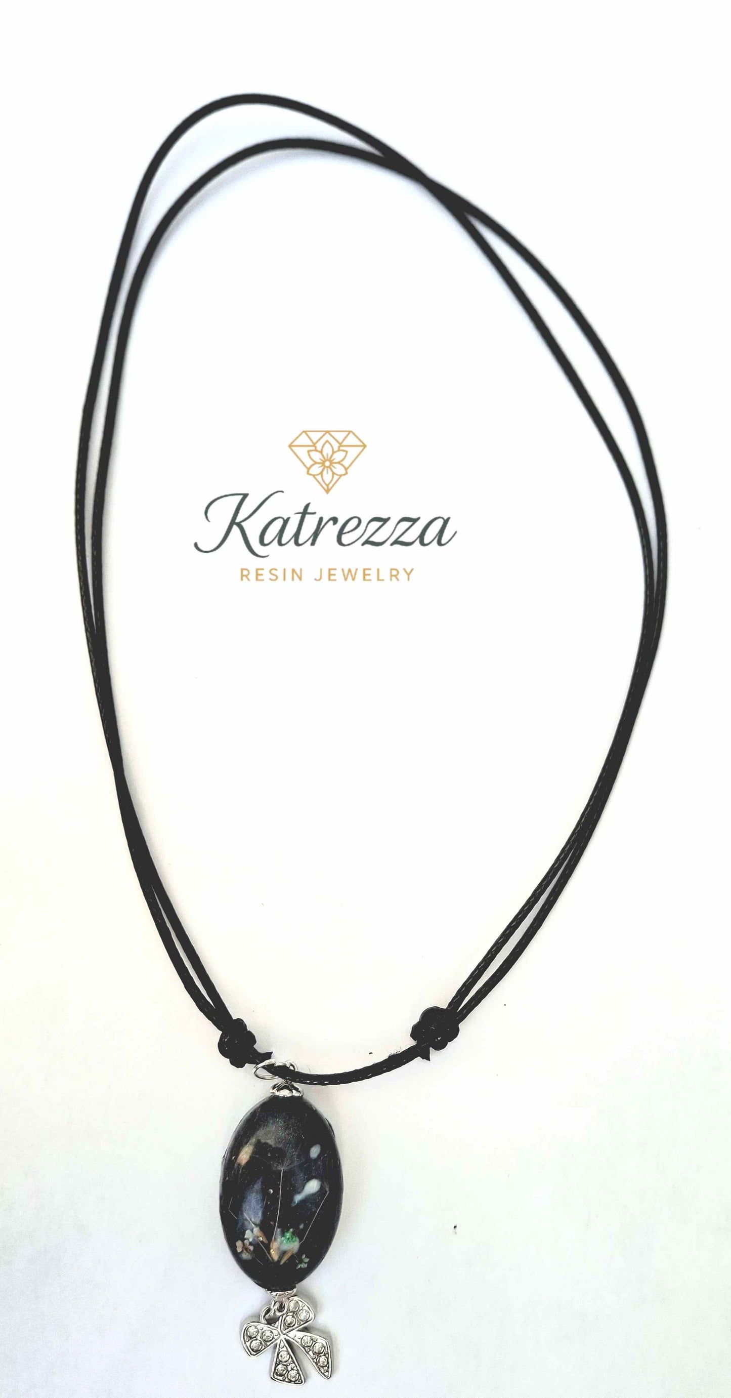 Adjustable black cord necklace with oval resin pendant containing Queen Anne’s Lace and dandelion seeds, and silver bow charm – Katrezza jewelry.