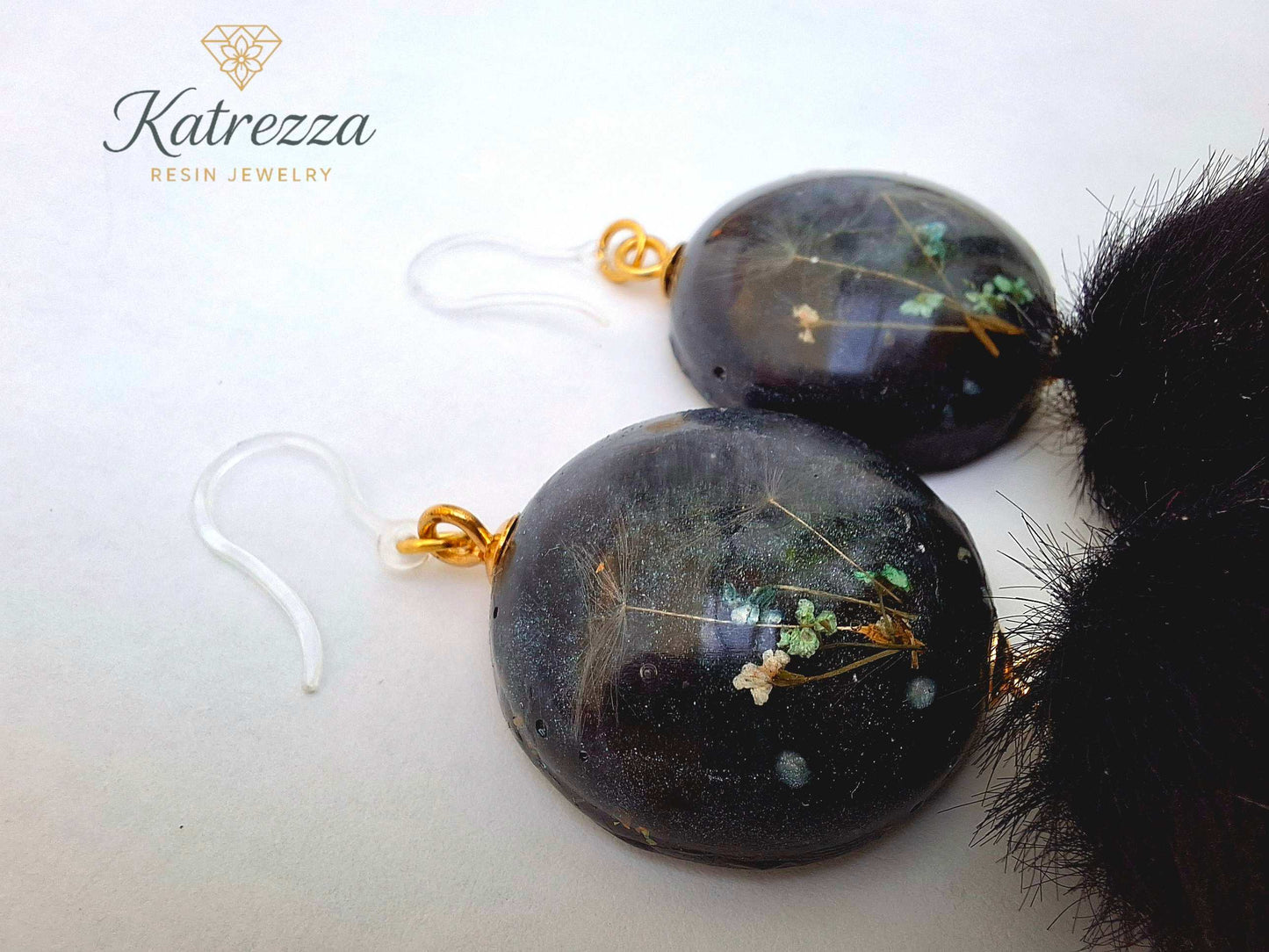 “Round resin earrings with real flowers and dandelion seeds in black resin, finished with fluffy black pom-poms and hypoallergenic silicone hooks.”