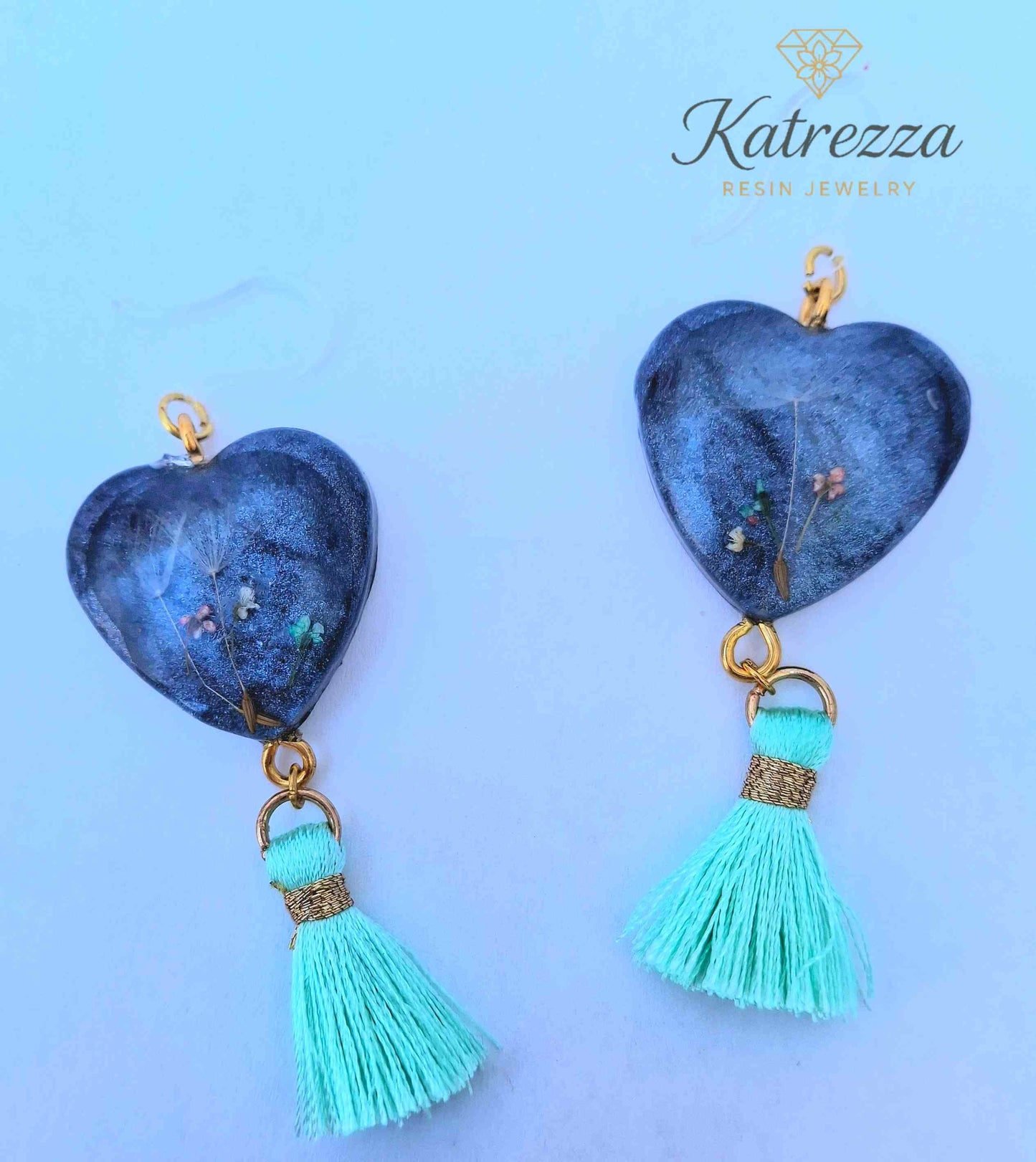 “Heart-shaped resin earrings with real flowers inside, black shimmer background, and mint tassels.”
