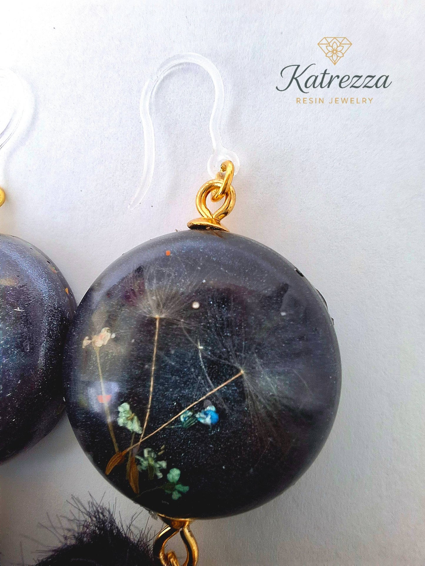 “Handmade black resin earrings with preserved flowers and dandelion seeds, accented with black pom-poms and gold details – hypoallergenic hooks.”