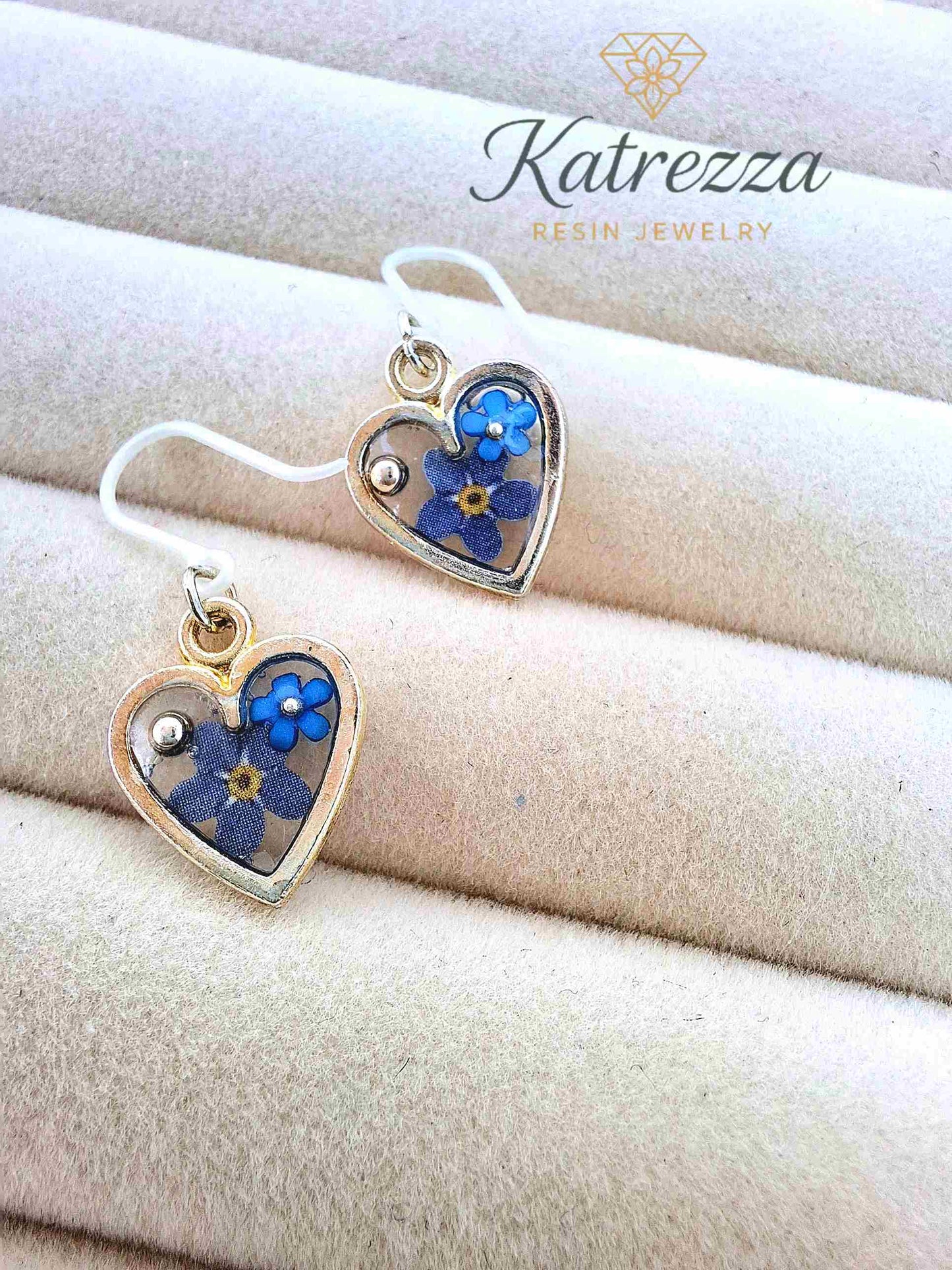 "Close-up of handmade silver heart-shaped charm earrings with resin interior and real blue pressed flowers, safe silicone hooks –