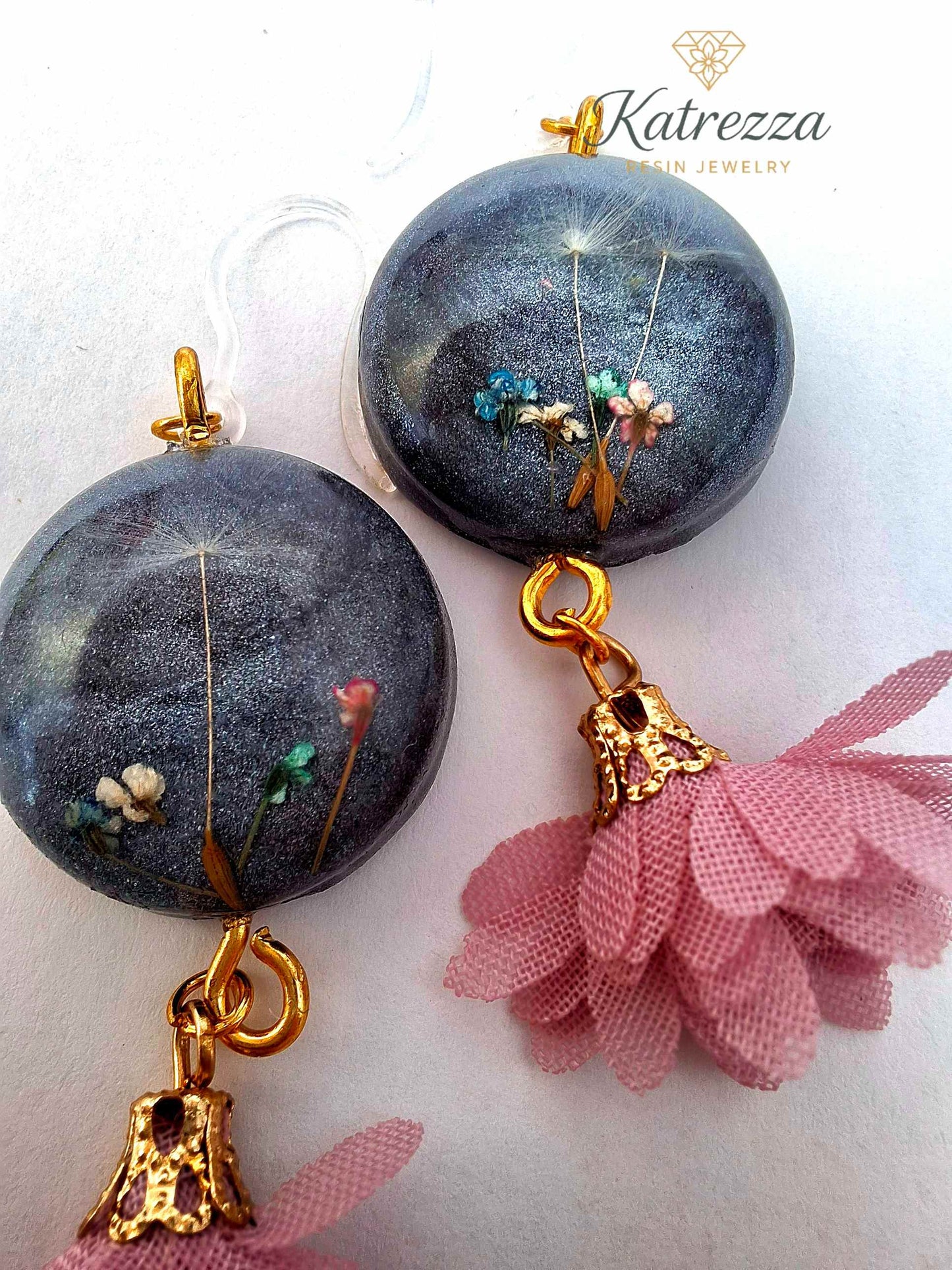 “Handmade 6.5 cm resin earrings with real flowers and dandelion seeds in black resin, gold accents, pink tassels, and silicone hypoallergenic hooks.”