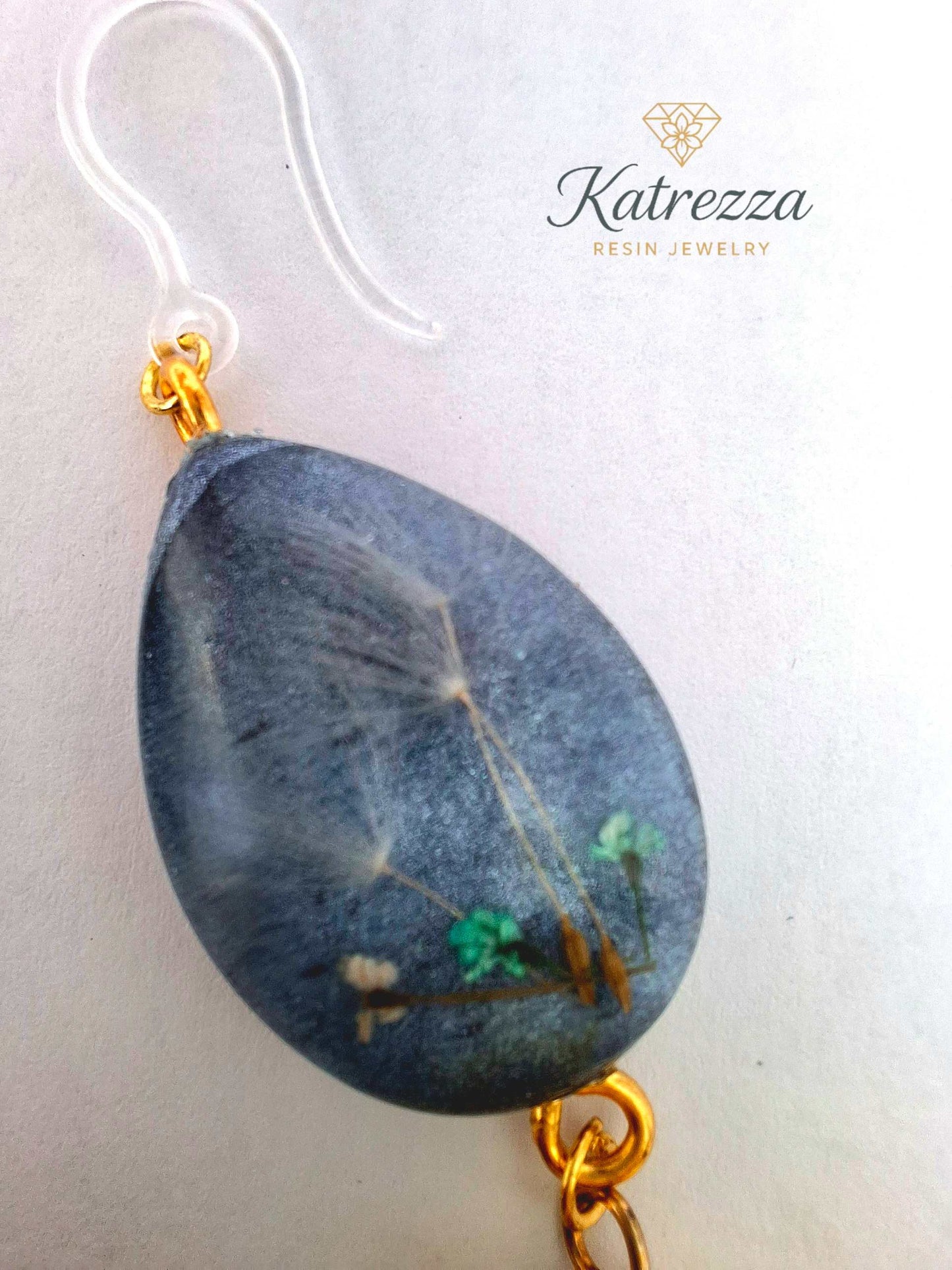 “Black resin teardrop earrings with tiny green flowers and dandelion seeds, finished with gold accents, yellow tassels, and hypoallergenic silicone hooks.”