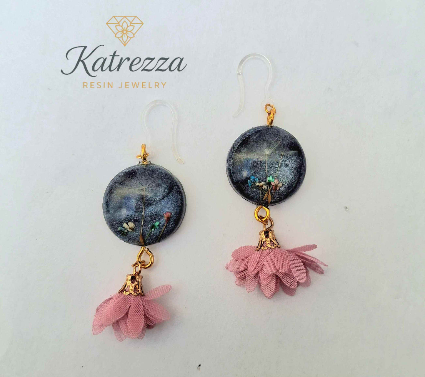 “Handmade 6.5 cm resin earrings with real flowers and dandelion seeds in black resin, gold accents, pink tassels, and silicone hypoallergenic hooks.”