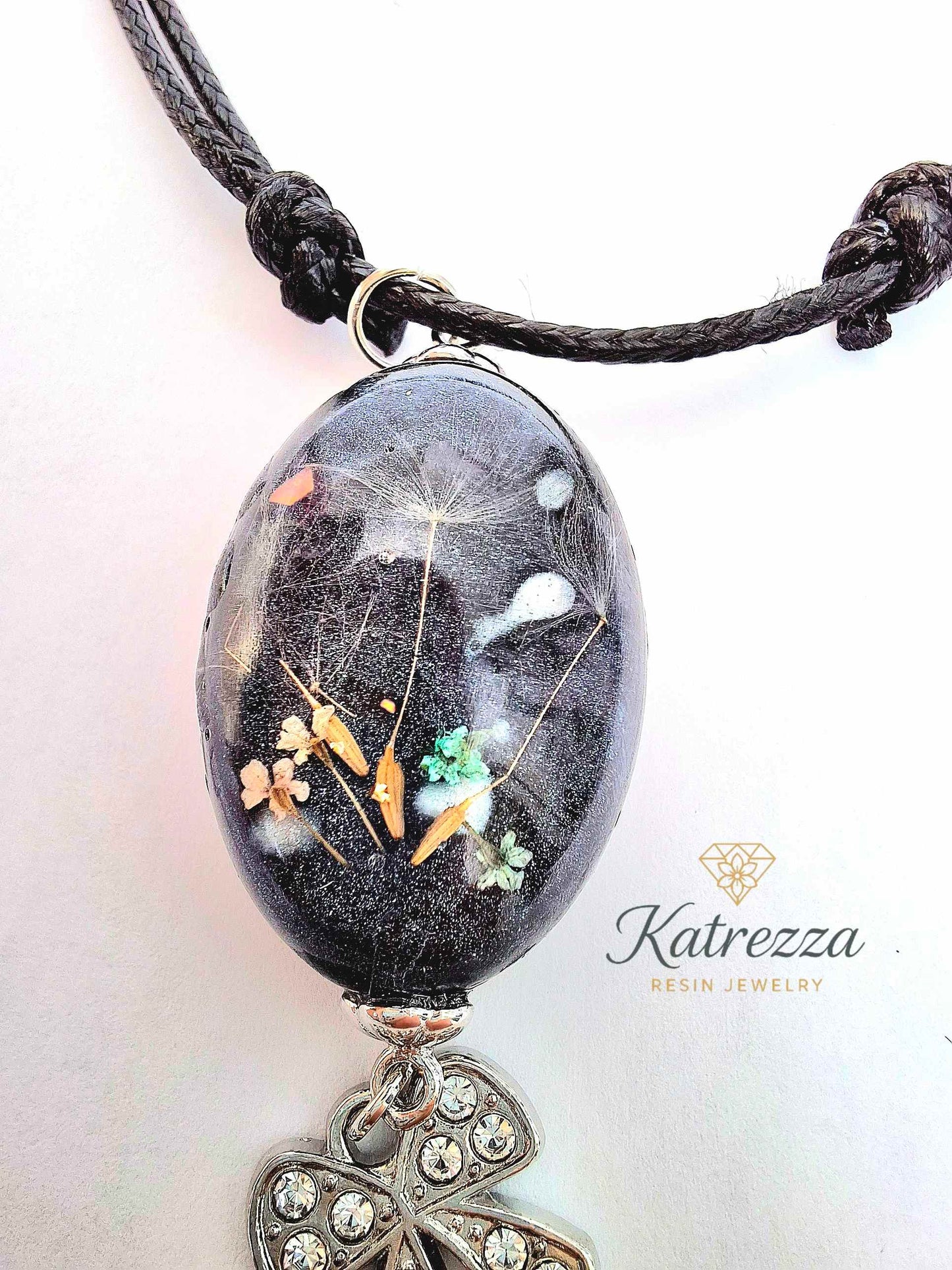 “Oval black resin pendant with real Queen Anne’s Lace flowers and dandelion seeds, finished with sparkling silver bow charm – Katrezza handmade jewelry.”