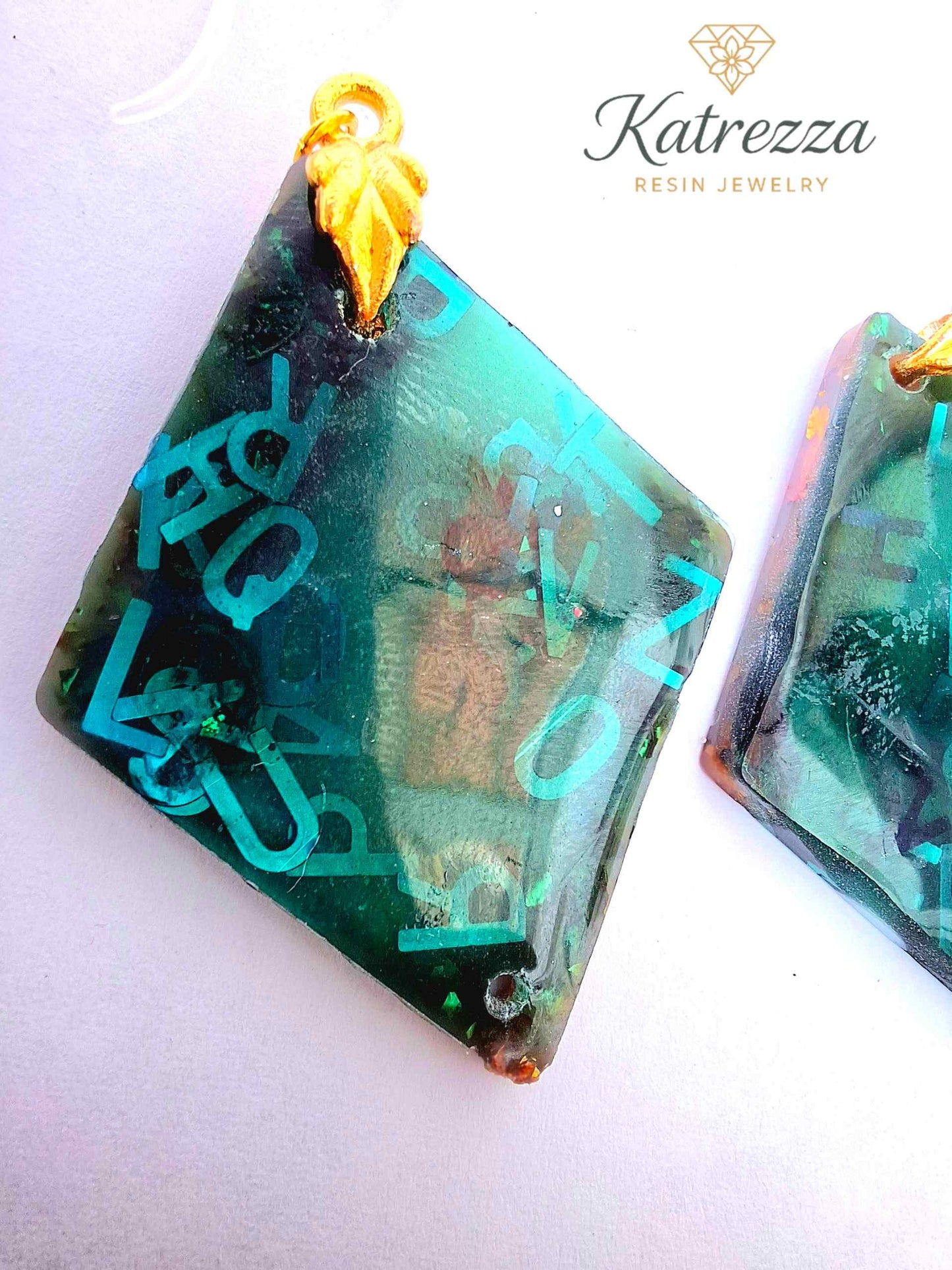 “Green resin earrings with shimmering blue letters and embedded details, geometric double-sided design.”