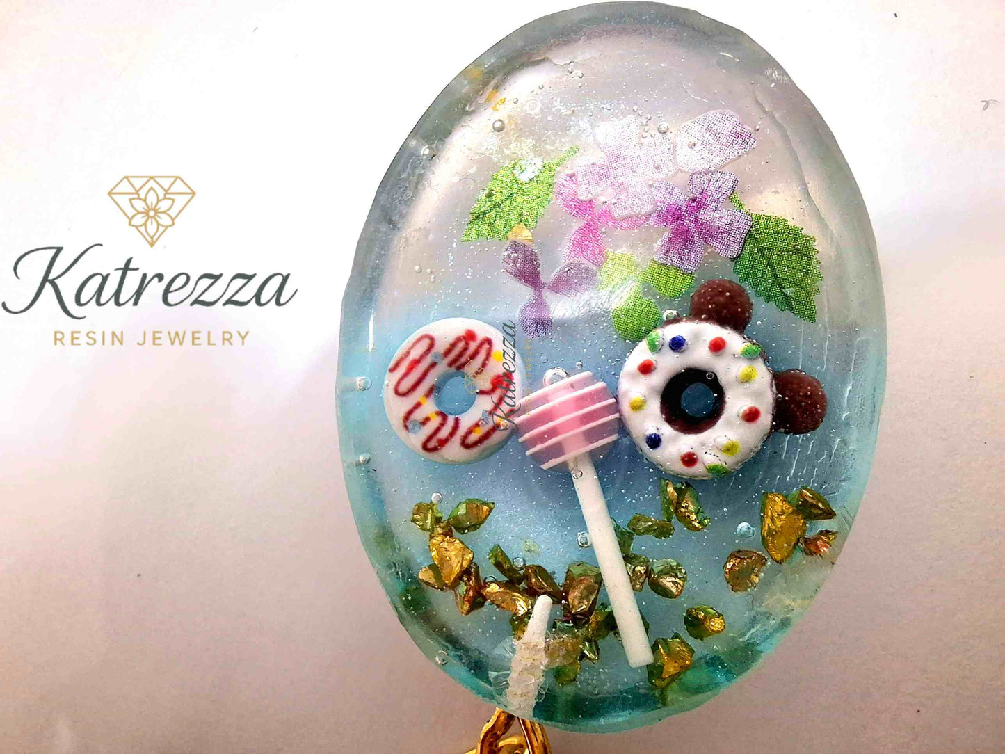 Side view of handmade oval resin keychain featuring donuts, lollipop, flowers, and golden flakes by Katrezza.