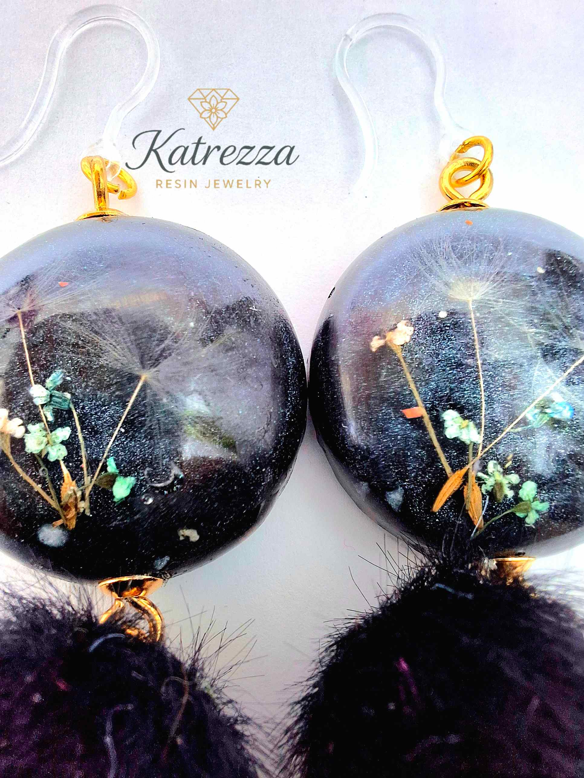 “Handmade black resin earrings with preserved flowers and dandelion seeds, accented with black pom-poms and gold details – hypoallergenic hooks.”