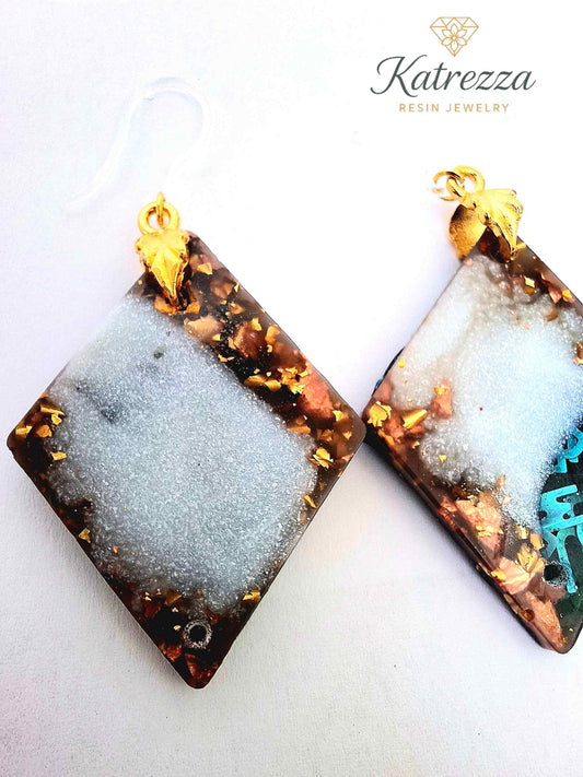 “Handmade resin earrings with gold flakes and white shimmer, diamond-shaped, 6 cm, hypoallergenic silicone hooks.”