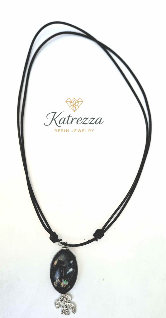 Adjustable black cord necklace with oval resin pendant containing Queen Anne’s Lace and dandelion seeds, and silver bow charm – Katrezza jewelry.
