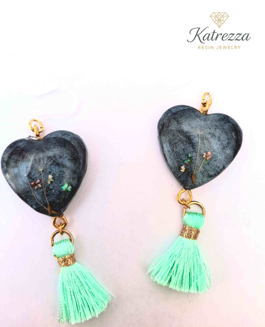 “Heart-shaped resin earrings with real flowers inside, black shimmer background, and mint tassels.”