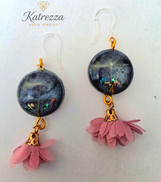 “Pair of handmade black resin earrings with real flowers, dandelion seeds, gold details, pink tassels, and silicone hypoallergenic ear hooks.”