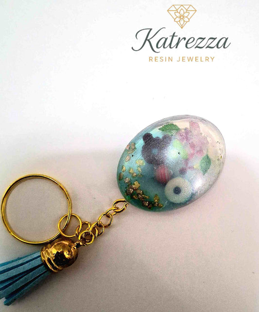 Handmade oval resin keychain with donuts, lollipop, purple flowers, and gold flakes by Katrezza Resin Jewelry.